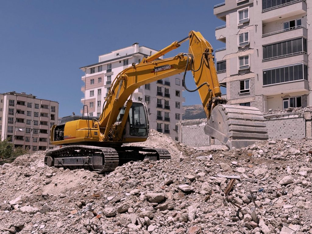 Yellow excavator working on urban construction site amidst rubble and buildings in Kahramanmaraş, Türkiye.